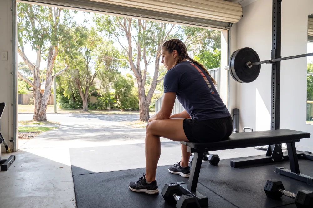 A woman resting on a bench between sets in a garage gym, wearing the Long Run Training Tee